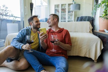 İki adam rahat oturma odalarının zemininde oturmuş kırmızı şarabı yudumlarken ve akıllı telefondan bakıp kahkahalarını ve gülümsemelerini paylaşırken birbirlerinin dostluğunun tadını çıkarıyorlar.