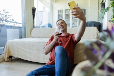Gülümseyen adam kanepenin yanında oturmuş, şık, modern bir oturma odasında akıllı telefonuyla bir bardak kırmızı şarabın tadını çıkarıyor.