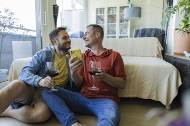 İki adam rahat oturma odalarının zemininde oturmuş kırmızı şarabı yudumlarken ve akıllı telefondan bakıp kahkahalarını ve gülümsemelerini paylaşırken birbirlerinin dostluğunun tadını çıkarıyorlar.