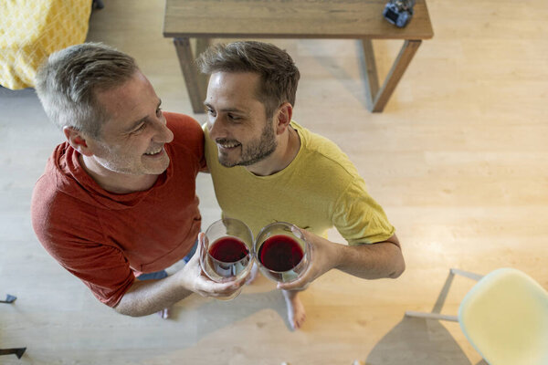 Двое мужчин обнимаются и улыбаются, держа бокалы красного вина, празднуя свою любовь и связь в уютном комфорте своего дома, наслаждаясь радостным моментом вместе