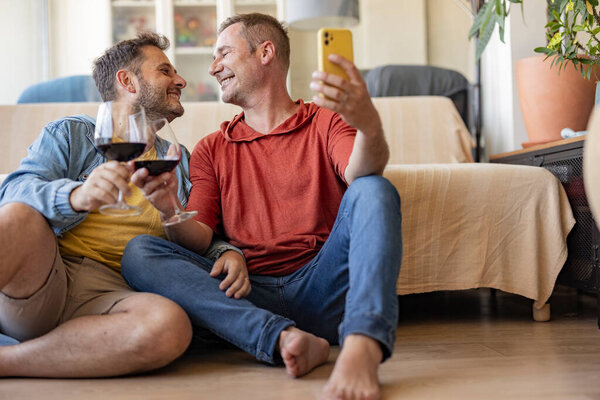 Двое мужчин сидят на полу своей современной гостиной, наслаждаясь бокалами вина, захватывая радостное селфи со своим смартфоном, излучая счастье и связь