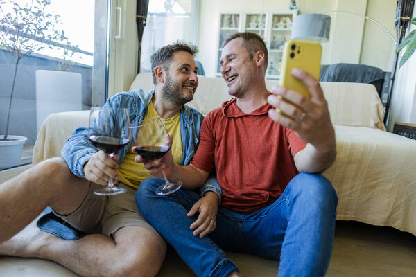 Двое мужчин сидят на полу, держа бокалы красного вина и делая селфи со смартфоном, наслаждаясь расслабляющим моментом вместе в их современной квартире