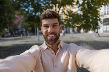 Şık saç kesimi ve küpeli mutlu genç adam Portekiz 'de halka açık bir parkta selfie çekiyor, gülümsüyor ve güneşli bir günün tadını çıkarıyor.