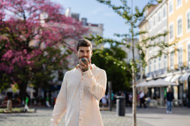 Güneşli Lizbon Meydanı 'nda yürüyen dövmeli genç bir adam, akıllı telefonuyla sesli bir mesaj gönderirken yakınlardaki çiçek açan bir ağacın canlı çiçeklerinin tadını çıkarıyor.