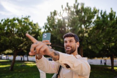 Mutlu genç yönetici kulaklık takıyor ve güneşli bir yaz gününde Lizbon, Portekiz 'deki bir parkta akıllı telefonuyla fotoğraf çekerken bir şeye işaret ediyor.