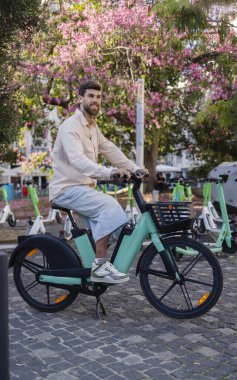 Güneşli bir günde arka planda park etmiş elektrikli scooterları olan, açık yeşil bir bisiklete binen, sıradan kıyafetler giyen, sakallı genç bir adam.