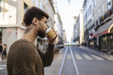 Boynunda dövme olan şık genç bir adam Portekiz 'de işlek bir şehir sokağının kaldırımında durup gününden biraz uzaklaşırken tek kullanımlık bir bardaktan sıcak bir içecek içiyor.