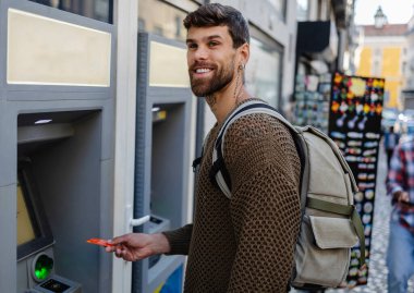 Genç erkek turist Portekiz 'deki bir ATM' den para çekiyor. Gülümsüyor ve kredi kartını makineye sokuyor.