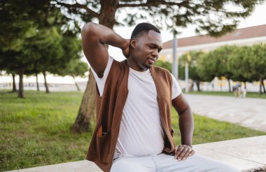 Tourist sitting on a low wall in a park in lisbon, portugal, grimacing in discomfort while touching his neck, clearly suffering from neck pain amidst the summer greenery clipart