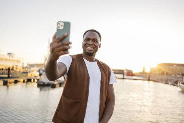 Portekiz, Lizbon 'da akıllı telefonuyla selfie çeken genç bir adam. Gün batımı, tagus nehrinin sularını yansıtıyor. Güzel bir altın ışık yaratıyor.
