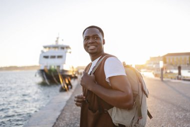 Günbatımında Lizbon Limanı 'nda arka planda feribot ile yürüyüp Portekiz' deki seyahat deneyiminin tadını çıkaran gündelik giysiler giyen genç siyahi bir adam.