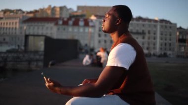 Portekiz 'de Tagus nehrinin kenarında oturan genç adam gün batımında canlı kentsel manzaranın tadını çıkarıyor. Altın saat ışığında dinleniyor, sakin atmosferi yakalıyor.