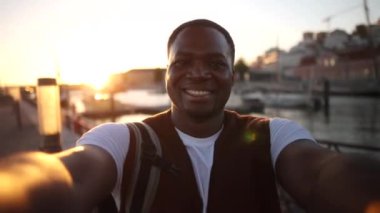 Mutlu Afrikalı Amerikalı turist sırt çantasıyla Aveiro, Portekiz 'de selfie çekiyor. Arka planda renkli evleri ve botları var. Avrupa' da tatilin tadını çıkarıyorlar.