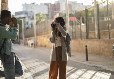 Sırt çantalı iki genç turist güneşli bir günün tadını çıkarıyorlar. Birbirlerine gülümserken klasik kameralarla gülümsüyorlar. Bu arada canlı şehir sokaklarını ve mimarisini keşfediyorlar.