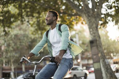 Bisikletle şehirde gezen, yaz havasının tadını çıkaran ve günlük kıyafetleri ve sırt çantasıyla başka yere bakan mutlu genç üniversite öğrencisi.