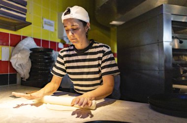 Çizgili tişört giyen kadın aşçı ve beyaz şapka ustalıkla pizza hamurunu yuvarlıyor. Londra 'nın hareketli bir restoranının mutfağında un broşu takılı.