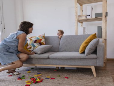 Büyükannesi kanepe yastığını kaldırırken torunu kanepenin arkasına saklanıp evde oynarken renkli ahşap yapı taşları halıya saçılmış.