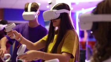 VR kulaklık takan ve kontrol cihazlarını elinde tutan bir grup genç oyuncu sanal gerçeklik oyun deneyimine dalmış, oyun oynamanın ve etkileşimli oyunların keyfini çıkarıyorlar.