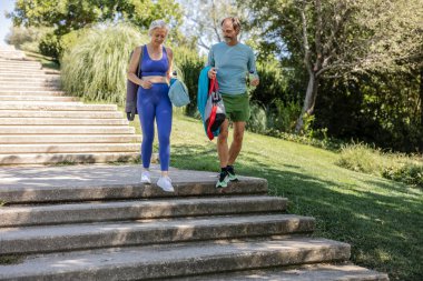 Üst düzey bir çift yoga dersinden sonra merdivenlerden aşağı iniyor. Paspaslarını, havlularını ve su şişelerini taşıyorlar. Sağlıklı bir yaşam tarzının keyfini çıkarıyorlar.