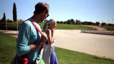 İki sporcu antrenmandan sonra parkta yürürler, spor çantaları taşırlar, spor ayakkabıları giyerler, sohbet ederler ve temiz hava alırlar.