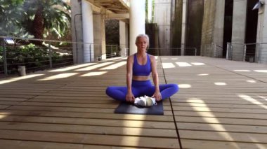 Olgun kadın, tahta zeminde kelebek yogası yapıyor, esnekliği ve farkındalığı artırıyor ve yaz günü huzurlu şehir parkı ortamının keyfini çıkarıyor.