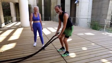 Üst düzey bir çift tahta güvertede savaş halatları egzersizi yapıyor. Yazın açık hava eğitimi sırasında güç, spor ve aktif yaşlanma gösteriyorlar.