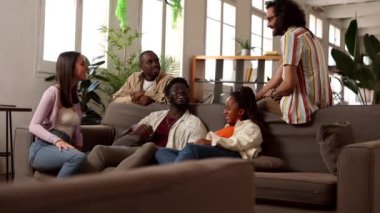 Çeşitli arkadaş grupları rahat bir anı paylaşıyor, birlikte oturuyor ve modern apartman ortamında rahat bir kanepede sohbet ediyorlar.