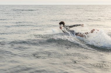 Young surfer paddling on a surfboard through the ocean waves at sunset, wearing a snug wetsuit and creating lively splashes as the golden hour light dances on the water clipart