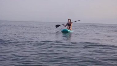 Sakin deniz yüzeyi alacakaranlık gökyüzünü yansıtıyor. Atletik kadın ayakta kürek çekiyor, aktif dinlenmenin ve deniz manzarasının tadını çıkarıyor.