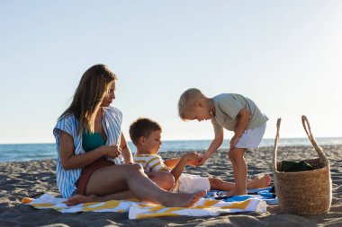 Anne ve iki oğul sahilde kaliteli vakit geçiriyorlar gün batımında çizgili havluyla oynuyorlar aile tatillerinin tadını çıkarıyorlar ve mutlu anılar yaratıyorlar.