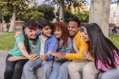 Beş neşeli çoklu etnik öğrenci akıllı telefonlarıyla selfie çekiyor, kampüs parkında oturuyor, sarılıyor, gülüyor, birlikte vakit geçiriyorlar.