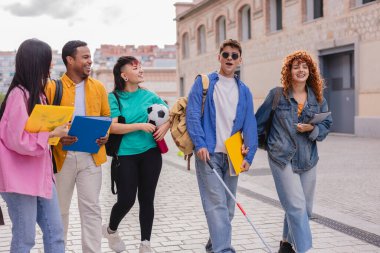 Kampüste birlikte yürüyen mutlu ve çeşitli üniversite öğrencilerinden oluşan bir grup, beyaz baston kullanan bir öğrenci, eğitime katılım ve destek gösteriyor.