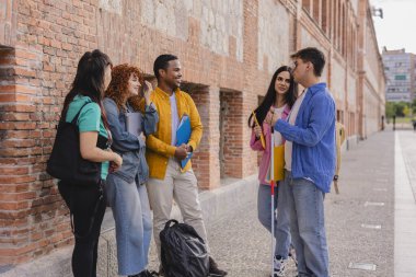 Çeşitli mutlu öğrenci grupları, bir üniversite binasının önünde beyaz baston kullanan bir öğrenciyle sohbet ediyorlar. Kampüste dostluk ve dostluk gösterisi yapıyorlar.