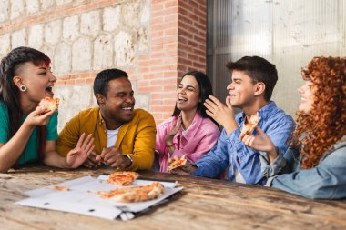 Bir grup neşeli çoklu etnik arkadaş birlikte pizza yemekten, eğlenmekten, gülmekten ve konuşmaktan zevk alıyorlar, pizzacıda ahşap bir masada oturuyorlar.