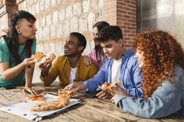 Bir grup mutlu çoklu etnik arkadaş kırsal bir ahşap masada pizza yerken, dilimleri paylaşırken ve iyi vakit geçirirken eğleniyorlar.
