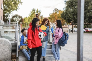 Çok ırklı bir grup öğrenci ellerinde not defterleri ve bir futbol topu ile güzel bir sonbahar gününde üniversitenin kampüs merdivenlerinde buluşup sohbet ediyorlar.