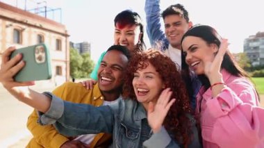Çeşitli genç arkadaşlar neşeli anları yakalıyorlar, güneşli bir günde dışarıda toplanırken gülümserken ve akıllı telefonlara el sallarken selfie çekiyorlar.