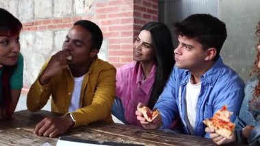 Genç arkadaşlar pizza dilimlerini paylaşıyorlar, gündelik sohbetleri ve kahkahaları seviyorlar, tuğla duvarın yanındaki ahşap masada otururken sıcak bir bağ yaratıyorlar.