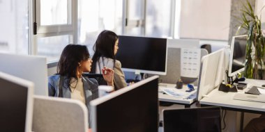 İki iş kadını modern bir ofiste bilgisayarlar üzerinde çalışıyor, ortak ve üretken bir ortamda doğal ışık ile görevlerine odaklanmış durumdalar.