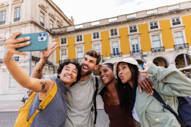 Dört mutlu çoklu etnik arkadaş grubu, Portekiz 'de sarı bir tarihi binanın önünde Lizbon' u ziyaret ederken akıllı bir telefonla selfie çekiyorlar.