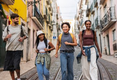 Sırt çantalı dört mutlu turist Lizbon 'da bir kaldırım taşı caddesinde yürüyor, tatillerinin tadını çıkarıyor ve şehrin tarihi alfama bölgesini keşfediyorlar.