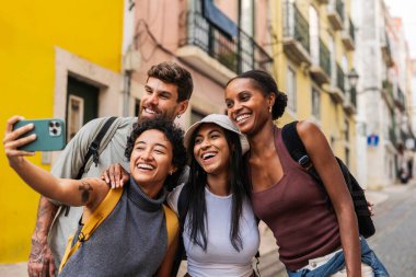 Lizbon, Portekiz 'in pitoresk şehrinde canlı bir yaz gününün tadını çıkarırken akıllı telefonlarıyla gülümseyen ve selfie çeken bir grup etnik arkadaş.