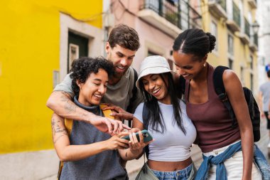 Sırt çantalı dört neşeli genç turist Lizbon, Portekiz 'de güneşli bir günün tadını çıkarıyorlar. Birlikte yürürken gülüyorlar ve akıllı telefonlarını kullanıyorlar. Keyif ve macera anları yakalıyorlar.