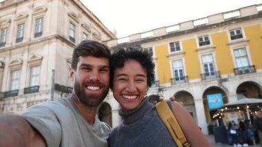 Lisbon 'un ticaret meydanında selfie çeken iki genç turist farklı mutluluk ifadeleri sergiliyor ve bazı önemli noktalara işaret ediyor.