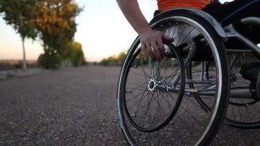 Tekerlekli sandalye kullanıcısı tekerlekli sandalyeye el bisikleti takıyor, gün batımında parktaki kaldırımlı yolda ilerliyor, adaptif spor ekipmanlarını ve aktif yaşam tarzını gösteriyor