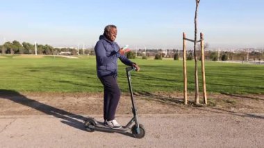 Yaşlı Afro-Amerikalı adam güneşli bir parkta elektrikli scooterıyla gezmekten keyif alır. Akıllı telefonunu navigasyon ve iletişim için kullanır.