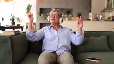 Kablosuz kulaklık takan mutlu emekli kadın oturma odasında rahat bir koltukta oturuyor, müzik dinliyor, şarkı söylüyor ve dans ediyor, boş zamanlarının tadını çıkarıyor.
