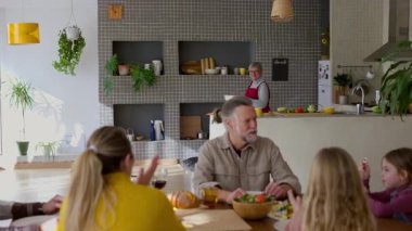 Çok nesilli aile yemek masasının etrafında toplanıyor, neşeli bir şekilde büyükanneyi alkışlıyor, ev yapımı yemekleri parlak, modern mutfak ortamında servis ediyor, sevgi ve bağlantıyı paylaşıyor.