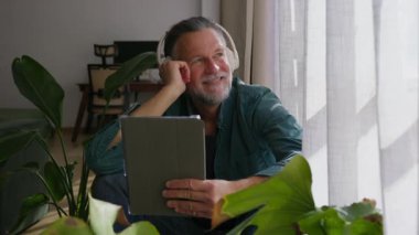 Kulaklık takan ve tablet tutan mutlu emekli adam, modern apartmanında pencerenin yanında otururken müziğin keyfini çıkarıyor.