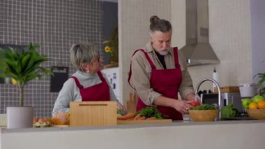 Üst sınıf çifti kırmızı önlük giyip besleyici yemek hazırlarken keyifli anları paylaşıyorlar. Taze sebzeleri yıkıyorum. Parlak modern mutfak alanında sevgi dolu gülümsemelerle sıcak bir şekilde iletişim kurmak.